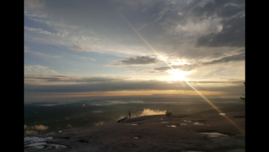 Sunrise view from the rocky summit of Stone Mountain, with a lone person standing near puddles in the foreground and golden light breaking through dramatic clouds over the distant landscape.