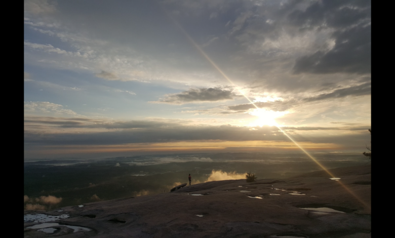 Sunrise view from the rocky summit of Stone Mountain, with a lone person standing near puddles in the foreground and golden light breaking through dramatic clouds over the distant landscape.